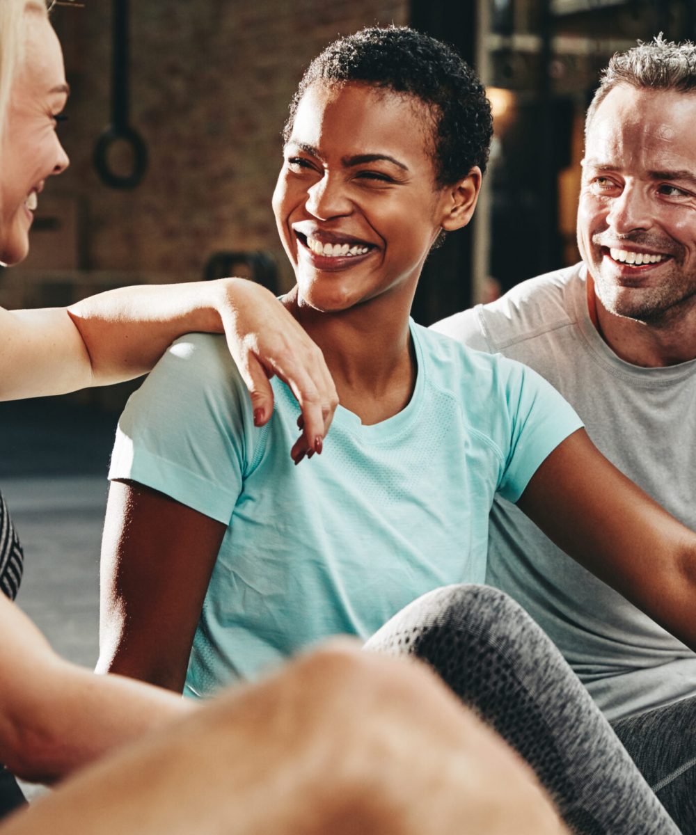Smiling group of diverse people in sportswear talking and laughing together while sitting on the floor after a gym workout class