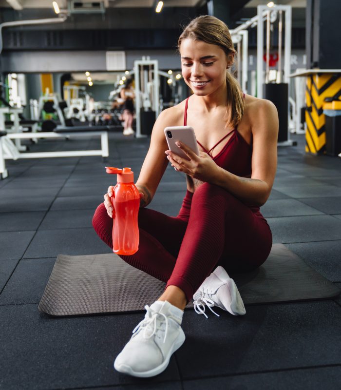 Photo of happy young woman fitness coach using mobile phone and drinking water in gym