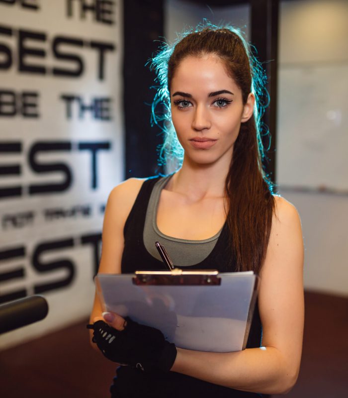 Portrait of beautiful female personal coach in gym. Girl holding results.